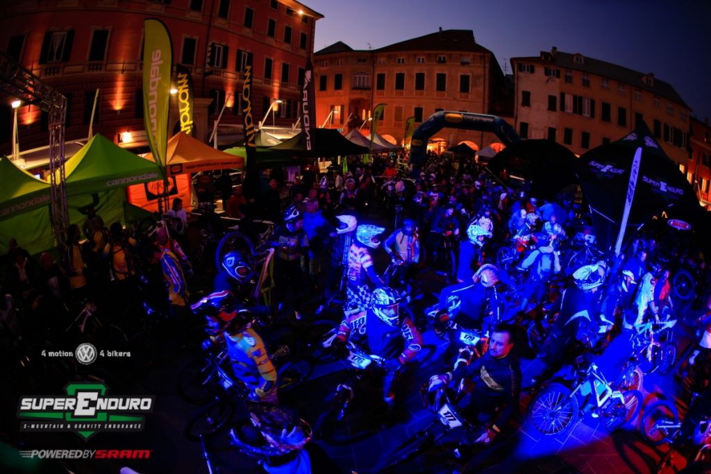Bikers in piazza illuminata durante evento mountain bike notturno.