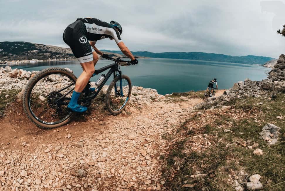Escursionisti in mountain bike su sentiero sconnesso con lago e montagne sullo sfondo.