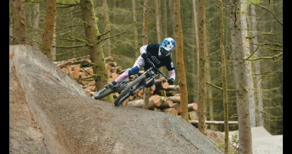 Pilota MTB in discesa su sentiero nel bosco con alberi e tronchi sullo sfondo.