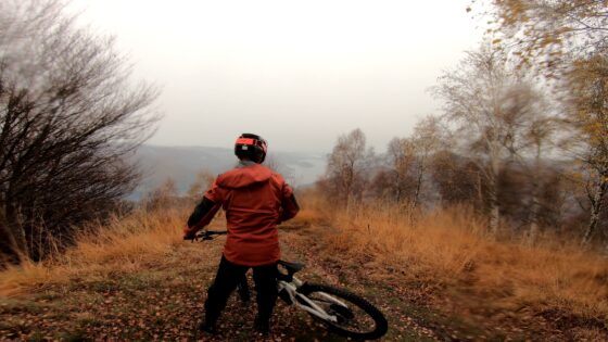 Sentiero di montagna autunnale con mountain bike e ciclista con casco rosso, paesaggio misto di verdi e arancioni.