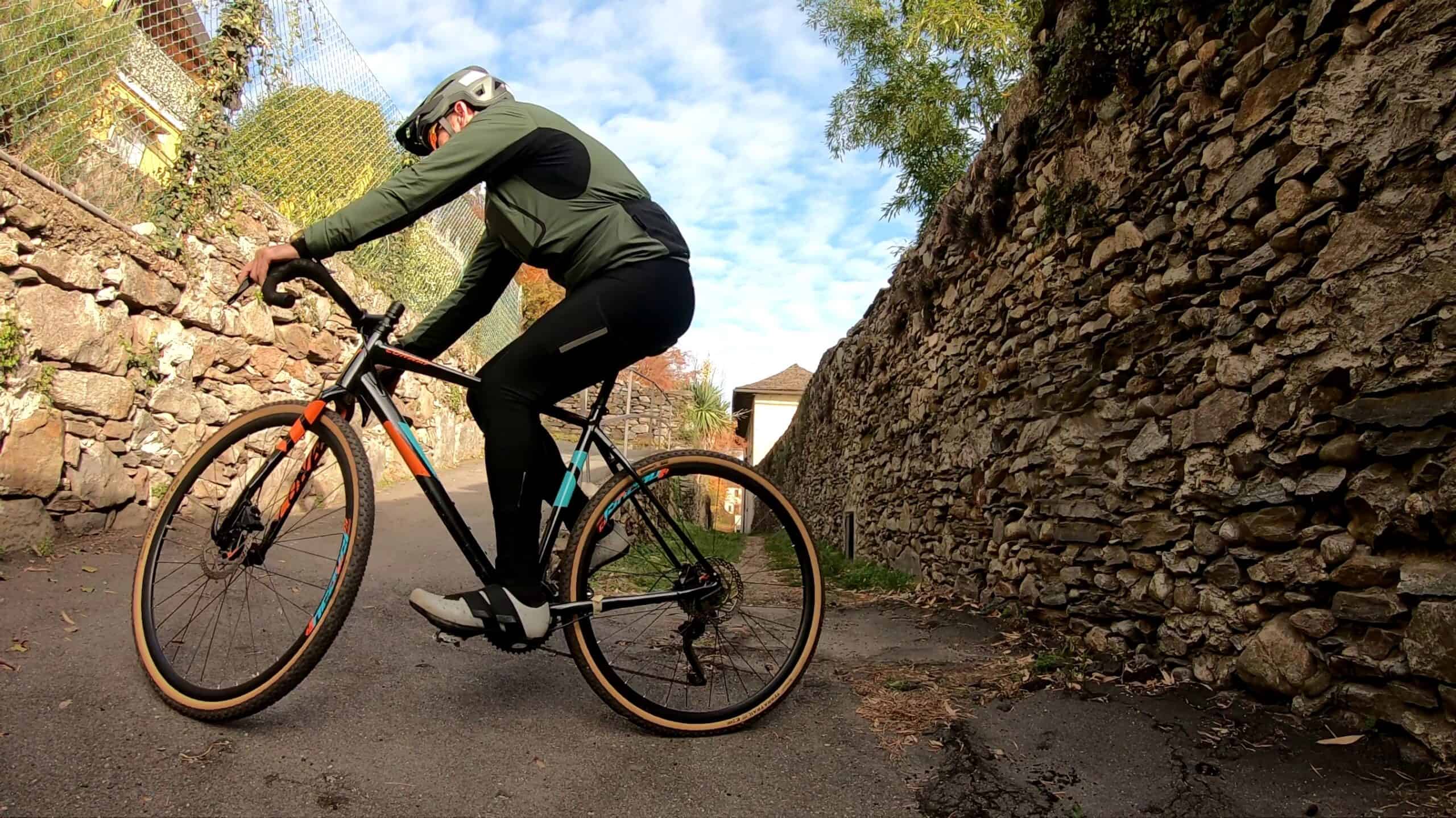 Telaio da mountain bike con muretti in pietra sullo sfondo, ciclista pronto a partire, abbigliamento tecnico e cielo azzurro.
