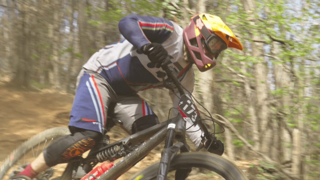 Pilota di mountain bike in azione su sentiero sterrato tra gli alberi.