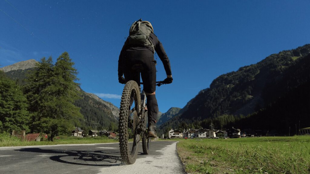 Adatto a meno di 100 caratteri: Escursionista in mountain bike su strada panoramica tra montagne verdi e azzurre.