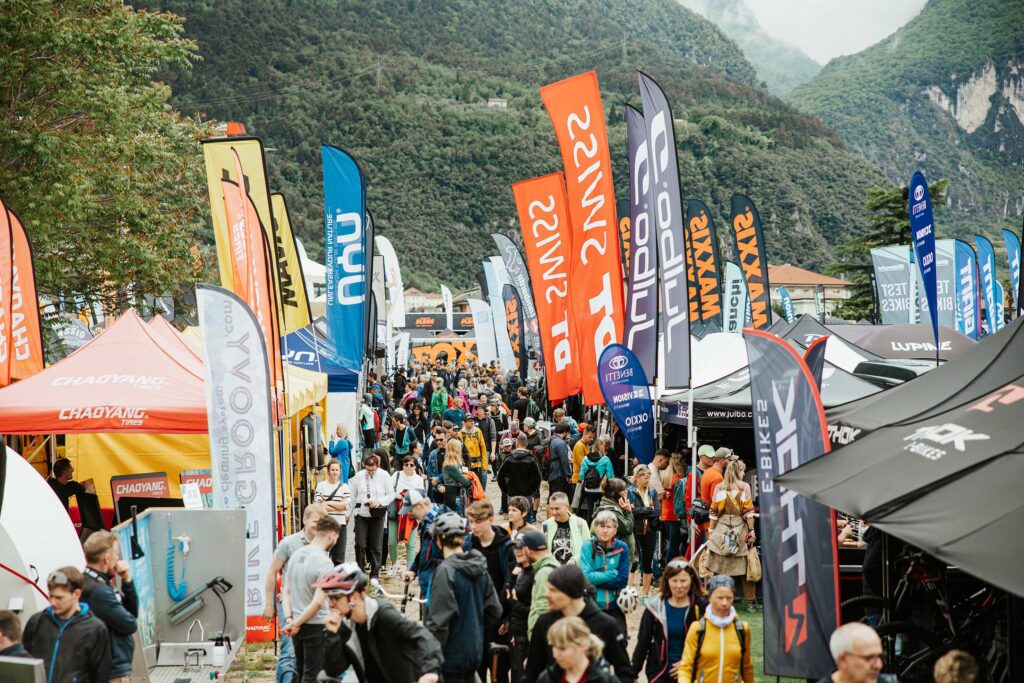 Folla numerosa a una fiera di mountain bike con molte bandiere di marche e stand all'aperto in una valle montuosa.
