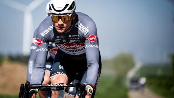 Ciclista in gara con caschetto e occhiali, su strada con vento e campagna alle spalle.