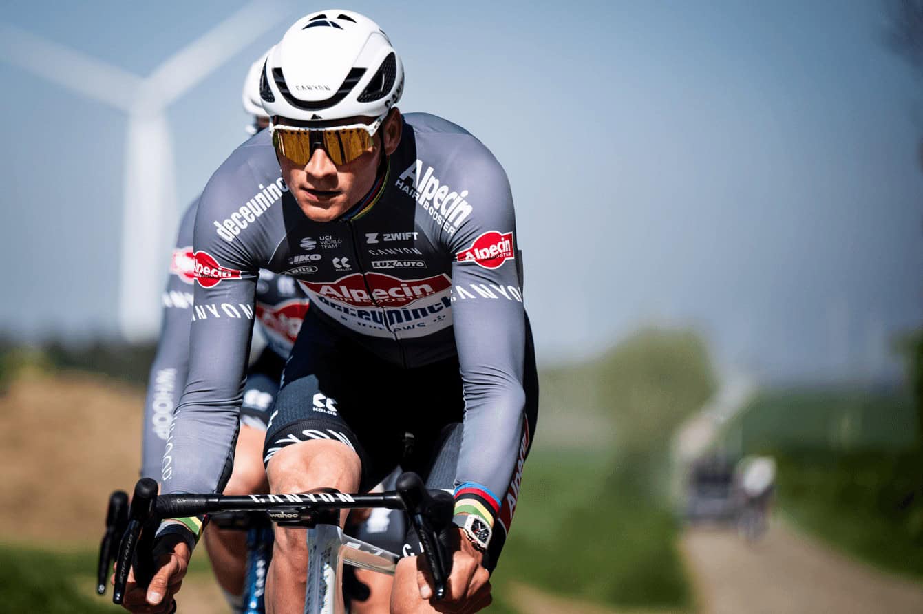 Ciclista in gara con caschetto e occhiali, su strada con vento e campagna alle spalle.