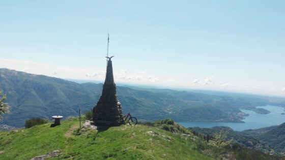 2600x1450 mountain bike with view of lake and hills in background, iconic monument at summit, bright sunny day, outdoor adventure, scenic landscape, cycling destination.