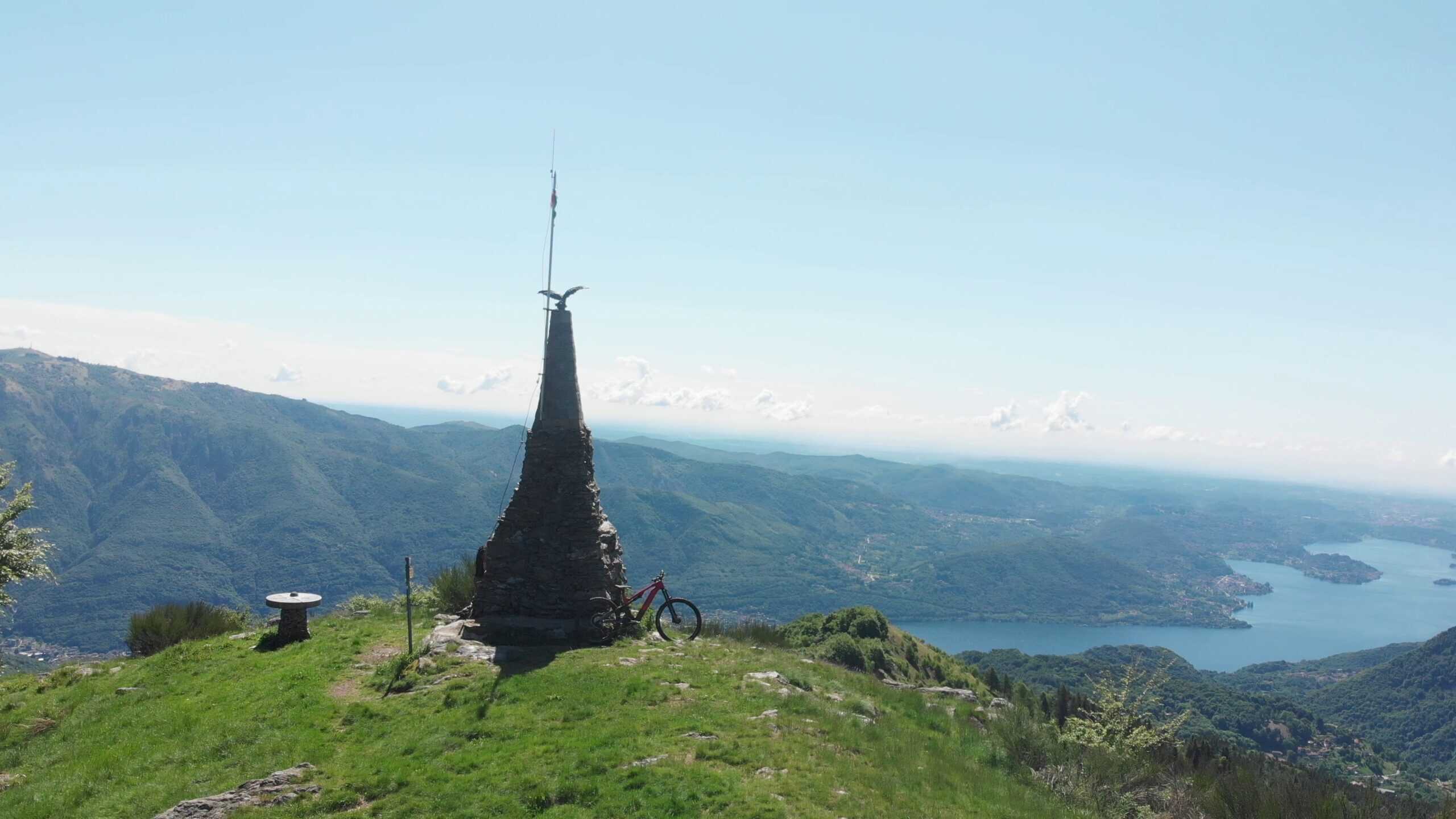 2600x1450 mountain bike with view of lake and hills in background, iconic monument at summit, bright sunny day, outdoor adventure, scenic landscape, cycling destination.