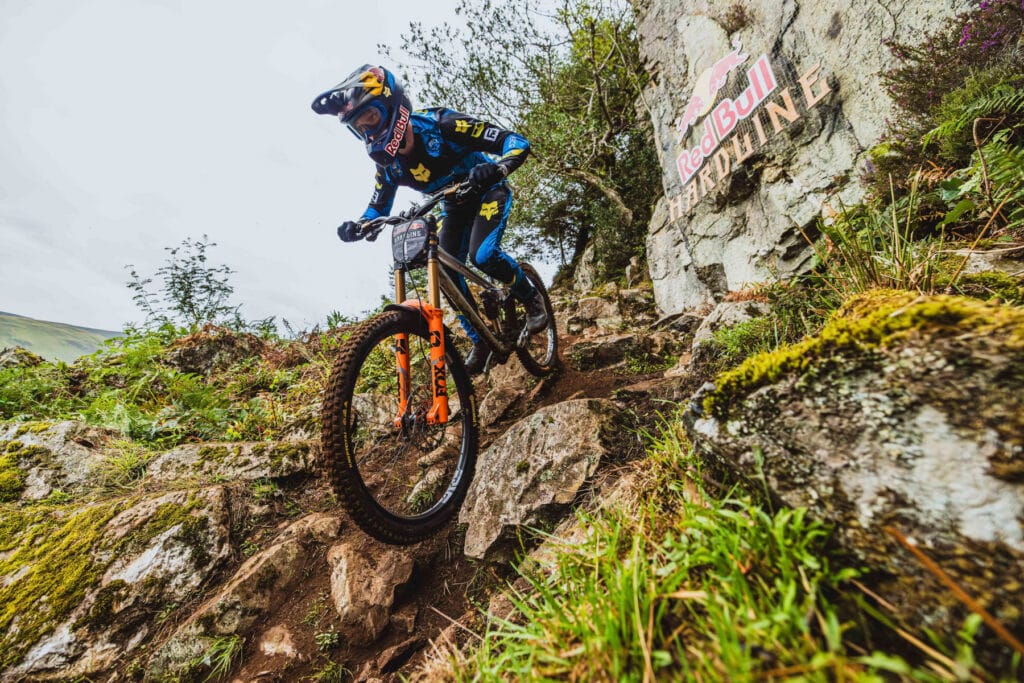 Corridore MTB in discesa su un sentiero roccioso in natura, con logo Red Bull in background.