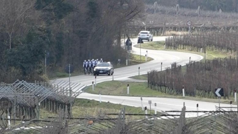 Auto della polizia in gara sulla strada di campagna, gruppi di ciclisti in sosta, paesaggio rurale con vigneti.