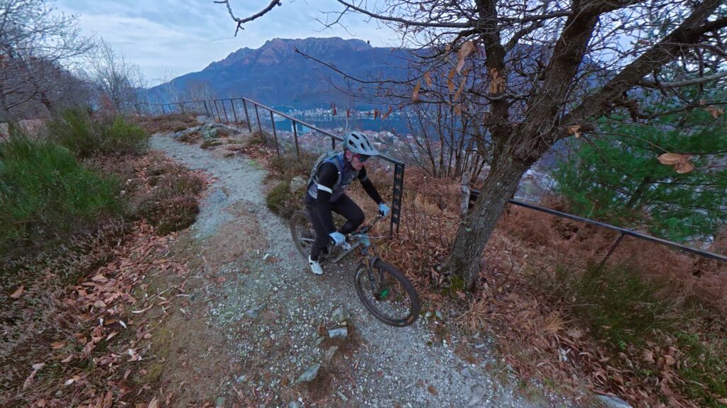 Salto in mountain bike lungo sentiero sterrato tra gli alberi e montagne sullo sfondo.
