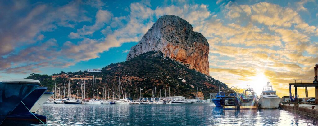 Montagna con barche in porto al tramonto, paesaggio naturale, cielo nuvoloso, vela e mare.