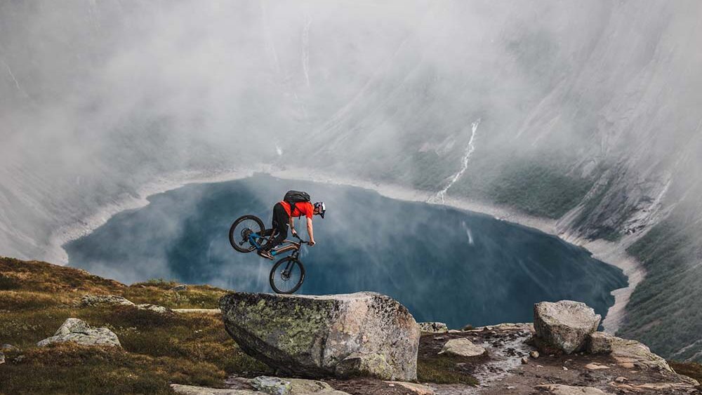 Acrobazia in mountain bike su roccia sopra un laghetto circondato da pareti di pietra.