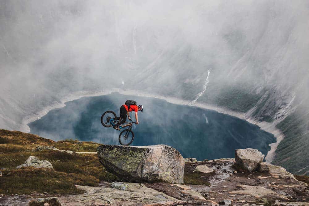 Acrobazia in mountain bike su roccia sopra un laghetto circondato da pareti di pietra.